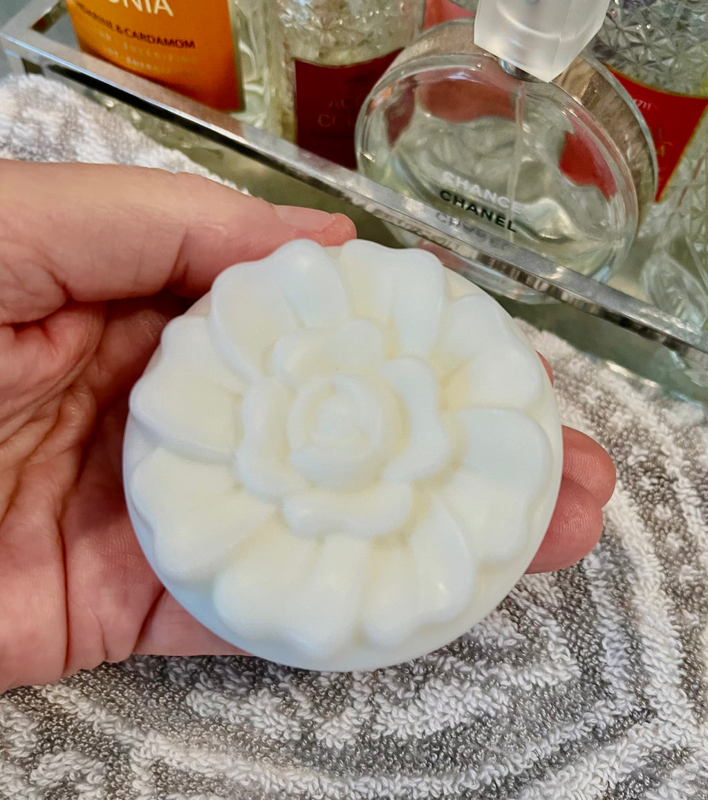 Hand holding a white floral lotion bar with blurred perfume bottles in the background