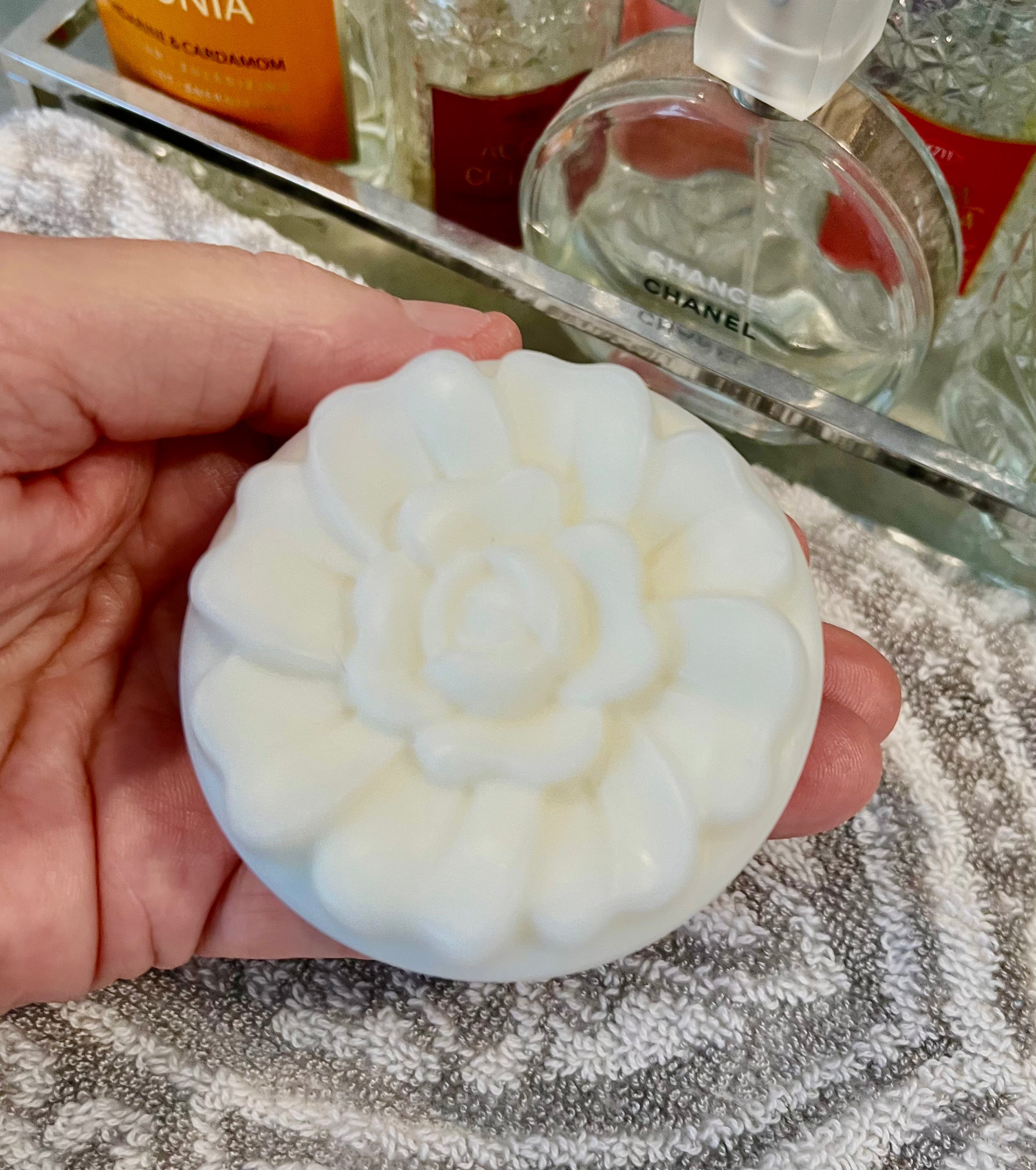 Hand holding a white floral lotion bar with blurred perfume bottles in the background