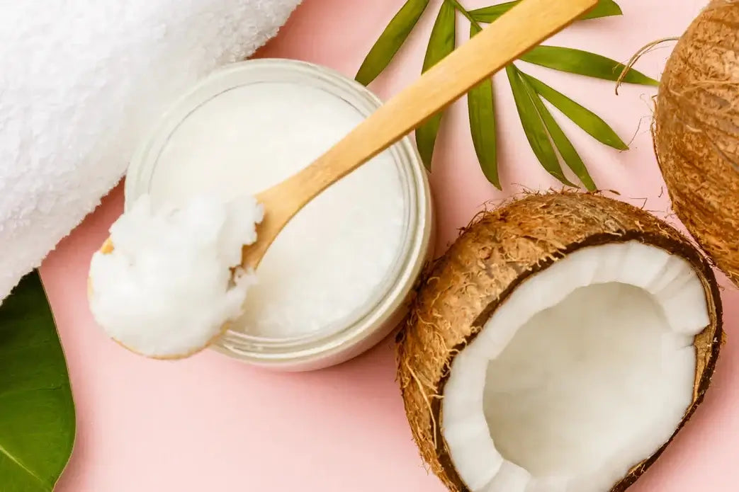 Coconut oil in a jar with a wooden spoon, half-coconut, and green leaves on a pink background